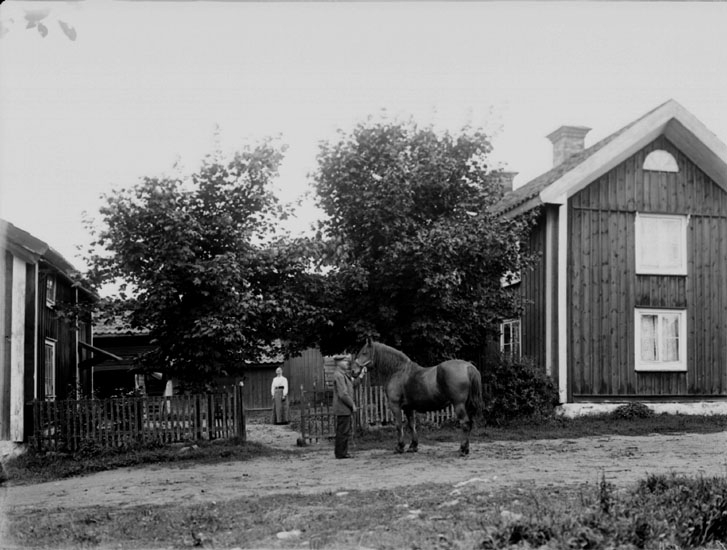 Byggnader, en man med häst, en kvinna.
Karl Erik Hansson