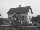 En och en halvvånings bostadshus med plåttak.
Grupp 9 personer framför huset.
K.G. Storm