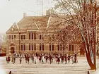 Trädgårdsskolan, tvåvånings skolbyggnad i tegel, skolbarn med lärarinna på skolgården.
Fröken Sofia Andersson