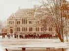 Trädgårdsskolan, tvåvånings skolbyggnad i tegel, skolbarn med lärarinna på skolgården.
Fröken Sofia Andersson