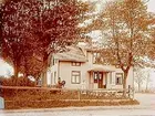 En och en halvplans bostadshus, med frontespis och förstubro.
3 personer framför huset.
K.A. Thulin