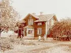 En och en halvplans bostadshus med frontespis och glasad veranda.
En vuxen och två pojkar framför huset.
Änkefru Anna Lovisa Olsson