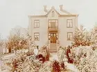 Tvåvånings bostadshus med frontespis och balkong, reveterat.
6 personer i trädgården framför huset.
Gustaf Engvall