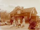 En och en halvvånings bostadshus med takhuv, förstebro och förstuga.
7 personer och en hund framför huset.
R. Bergström