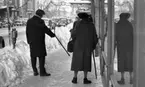 Halkiga pensionärer 18 december 1965

Två pensionärer står på trottoaren en vinterdag. Det är en man och en kvinna. Mannen pekar med sin käpp på en isfläck på marken och kvinnan tittar på stödjande sig på sin käpp. Till vänster om mannen syns en parkeringsautomat. En stor uppskottad snöhög löper vid sidan längs med trottoarkanten. Parkeringsautomater sticker upp ur snöhögen. Framför de två pensionärerna längre fram på gatan ligger Handelsbanken. På gatan står bilar parkerade längs med trottoarkanten. Uppe i luften över gatan hänger juldekorationer.