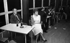 Minnenas 23 maj 1966
Vid ett bord sitter en kvinna i vit ärmlös blus och veckad kjol samt med en cigarett i höger hand. Bredvid henne sitter en man i ljus kavaj, vit skjorta och svart slips. På bordet står två Coca- Colaflaskor med sugrör i, många sugrör, ett askfat med fimpar i samt en tändsticksask. I bakgrunden syns fler bord med andra kvinnor och män.