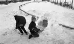 Snögrottor, 4 december 1965 

Man varnar för 'farliga snögrottor' i snöhögarna från snöplogarna. Två pojkar har skyfflat upp en hög med snö bakomstaketet runt gräsmattan. Nu håller den ene pojken på att gräva ur öppningen till grottan medan den andre skyfflar upp mer snö.