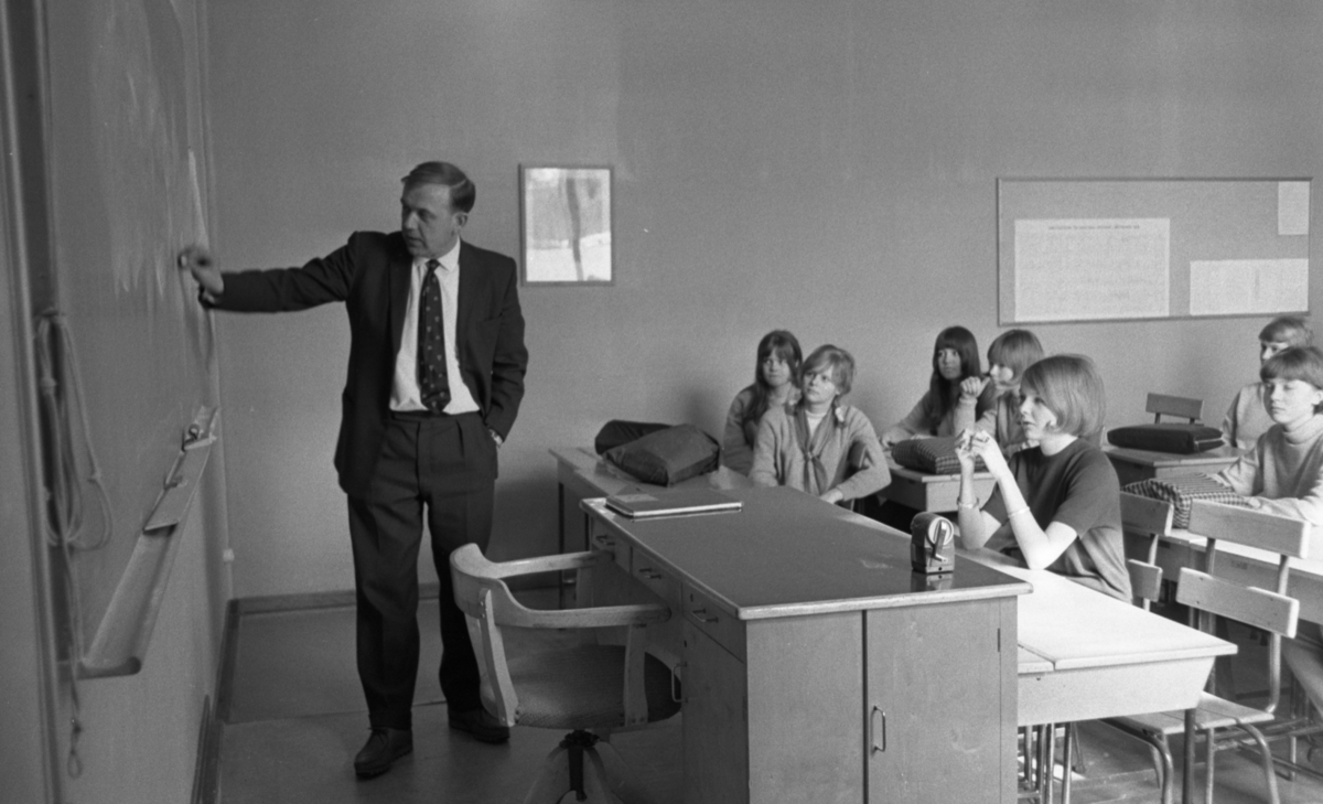 Engelsk lärare 6 april 1966

En lärare i engelska står framför svarta tavlan i klassrummet och skriver på den. I förgrunden står en kateder och en stol. Bakom läraren sitter elever i skolbänkar.















































































































or. Han går nedför en kort trappa utomhus.