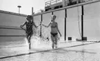 Hej Sommar 3 maj 1966

Två kvinnor - en  i baddräkt och en i bikini håller varandra i händerna och springer genom vattnet i den grunda bassängen i Gustavsviksbadet.








































































































































































































































































or. Han går nedför en kort trappa utomhus.