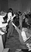 Popgala i Kumla, Farlig skolväg 24 augusti 1965

Ett rockband spelar på en scen i Kumla. En av medlemmarna har en gitarr i sina händer. En tonårsflicka ur publiken har klättrat upp på scenen och sitter på den. Hon sträcker ut sin högra hand mot gitarristen. En annan gitarrist syns i bakgrunden samt en sångare med en Sherlock Holmesinspirerad hatt på huvudet. En bandmedlem med tamburin i händerna syns längre bort i bakgrunden samt ett piano. Publik står nedanför scenen.