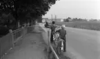 Popgala i Kumla, Farlig skolväg 24 augusti 1965

Två barn står med sina cyklar vid en trafikerad gata. En bil samt en buss har precis kört förbi dem. Byggnader syns i bakgrunden. Till vänster syns en grind och staket.