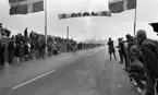 Cykel, 6 juni 1966

Ett antal cyklister cyklar in i mål under en tävling. Publik står på båda sidor om vägen och tittar på. Två svenska flaggor står uppställda vid var sin sida om vägen. Över vägen hänger en skylt med texten: 