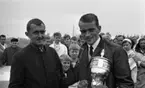 Cykel, 6 juni 1966

Två män under en cykeltävling. Mannen till höger håller i en prisbuckla samt blommor. Publik skymtar i bakgrunden.