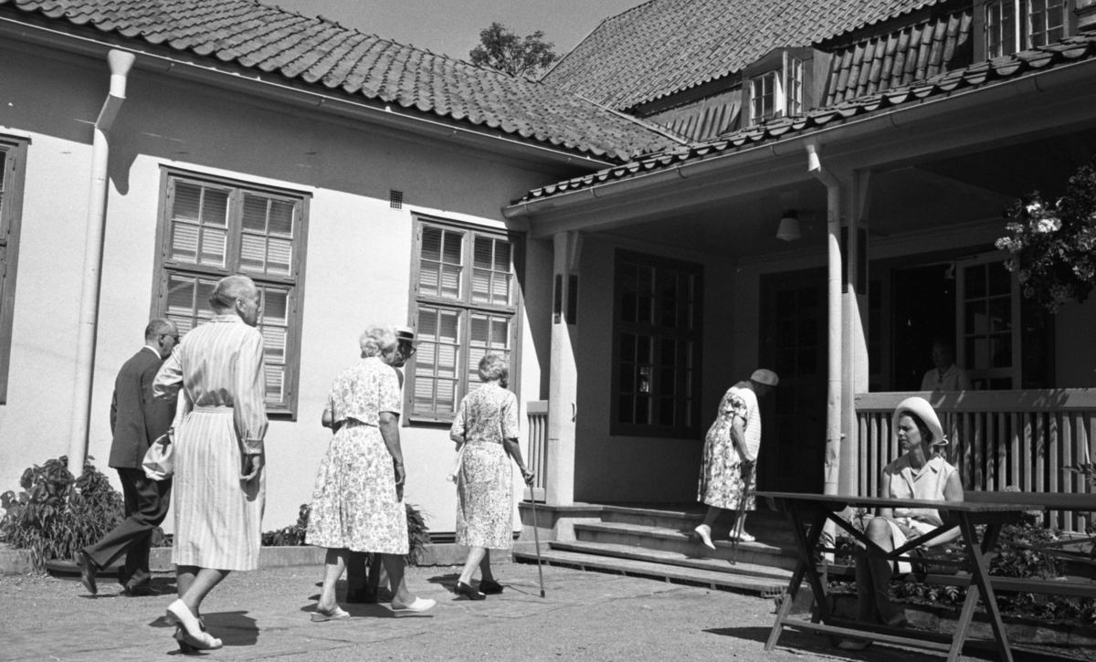 Loka brunn 22 juli 1966

Fyra äldre damer klädda i klänningar och två äldre herrar klädda i kostymer är på väg mot entrén på en av byggnaderna i kurorten Loka brunn. Till höger sitter en yngre kvinna vid ett bord. I dörröppningen till entrén står en annan kvinna.