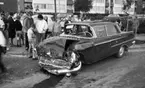 Krock Baronbacken 7 aug 1967. 
En mörk bil som varit med om en krock och har intryckt motorhuv står på gatan. Runtomkring står en massa människor. Byggnader syns i bakgrunden.