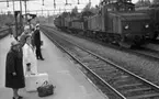 Frövi 2 juni 1967

Resenärer i olika åldrar står på perrongen på Frövi järnvägsstation. Två äldre damer står i förgrunden. Den första damen bär en ljus hatt, mörk kappa, mörka skor samt glasögon. Den andra damen är klädd i vit kappa. En liten mörk resväska med ett paraply i samt en stor vit resväska står på marken mellan damerna. En stins står i bakgrunden och gör honnör. Fler personer syns i bakgrunden. Två tåg syns längre bort.