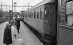 Frövi 2 juni 1967

Resenärer i olika åldrar står på perrongen på Frövi järnvägsstation. Två äldre damer står i förgrunden. Den första damen bär en ljus hatt, mörk kappa, mörka skor samt glasögon. Den andra damen är klädd i vit kappa. En liten mörk resväska med ett paraply i samt en stor vit resväska står på marken mellan damerna. En stins står i bakgrunden och viftar med en flagga. Ett står vid perrongen.