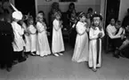 Luciafirandet 13 december 1966

Ett antal barn i sexårsåldern går i ett Luciatåg. Längst fram går en liten Lucia klädd i vitt linne med rött skärp om livet och med en Luciakrona med ljus på huvudet. Hon håller ihop sina händer. Efter henne kommer fyra små tärnor också i vita linnen och med glitterkransar på sina huvuden. Därefter kommer en stjärngosse med en Staffansstjärna i höger hand och strjärngossestrut på huvudet. Ytterligare små barn klädda i kockkläder, icke utklädda barn m. fl. går i tåget. I bakgrunden sitter publik: många mammor med små barn i famnen