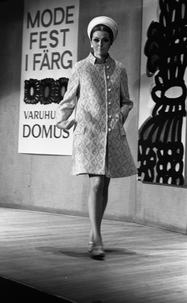 Modevisning, Görtz motor (Rep av oil) 6 april 1967

En fotomodell går på en scen under en modevisning. Hon är klädd i en kort, mönstrad ljus kappa, vita skor, vit hatt och har vita örhängen i öronen. Bakom henne syns en skylt på väggen där det står: "Modefest i färg varuhuset Domus."