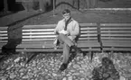 Wadköping (...?), 30 augusti 1968

En man sitter på en parkbänk i Wadköping. Han är med och ordnar eller repeterar en teaterföreställning. Han är klädd i beige rock, ljus skjorta, mörk slips, ljusa byxor, mörka strumpor och mörka skor. Han bär även glasögon samt håller några pappersark i sina händer. Två andra parkbänkar står på ömse sidor om honom. Byggnader syns i bakgrunden.