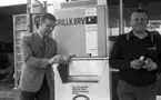 Korvätandet 30 augusti 1968

En man står och håller i en grillkorv som han precis har köpt i en automat. Han är klädd i ljus kavaj, ljus skjorta, mönstrad slips och mörka byxor. Bredvid honom står en man i mörk tröja och ljusa byxor. Han håller något i sina händer.