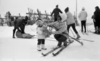 Ånnaboda 12 februari 1968

En liten flicka står i Ånnabodas skidbacke med en nalle i famnen och ropar något. Hon är klädd i vit mössa med mörk tofs på huvudet, mörk halsduk, vit jacka, mörka byxor och vita stövlar. Invid henne knäböjer en kvinna klädd i vit mössa, vitmönstrad jacka, svarta byxor och svarta pjäxor. Hon har skidor på fötterna och håller ett par stavar klämda mot vänstra sidan av kroppen med vänster arm. Fler personer samt ett ställ med skidor syns i bakgrunden. En person håller på och lastar något i en pulka.