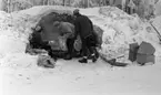 Ånnaboda, fotojobb (?), arbetarkommun 70 år 12 februari 1968

En kvinna och två tonårspojkar befinner sig ute i naturen i Ånnaboda. De befinner sig vid sidan om en snödriva med en utskjutande kant över. Det ser ungefär ut som en halv grotta. De har tak över sina huvuden. Kvinnan sitter ned och de unga tonårspojkarna står upp. Kvinnan är i färd med att ge en av pojkarna skräp som han lägger i en plastpåse. Alla tre är klädda i mössor, jackor, byxor och pjäxor. En annan person sitter delvis skymd i bakgrunden. Till höger ligger en liten pulka samt tre lådor på marken.