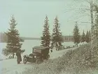 En man står vid sin bil på landsvägen nära en sjö. 
Bilen är en Oldsmobile modell 30 från 1923-24 med en 42 hästkrafters sexcylindrig motor. Den registrerades den 2:e april 1924 på fröken Jenny Larsson, Örebro, och fick registreringsnummer T99. År 1930 ägdes den fortfarande av Jenny Larsson.