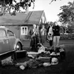 Familjegrupp fyra personer vid bilen. Bostadshus i bakgrunden.
Hallebys resa.