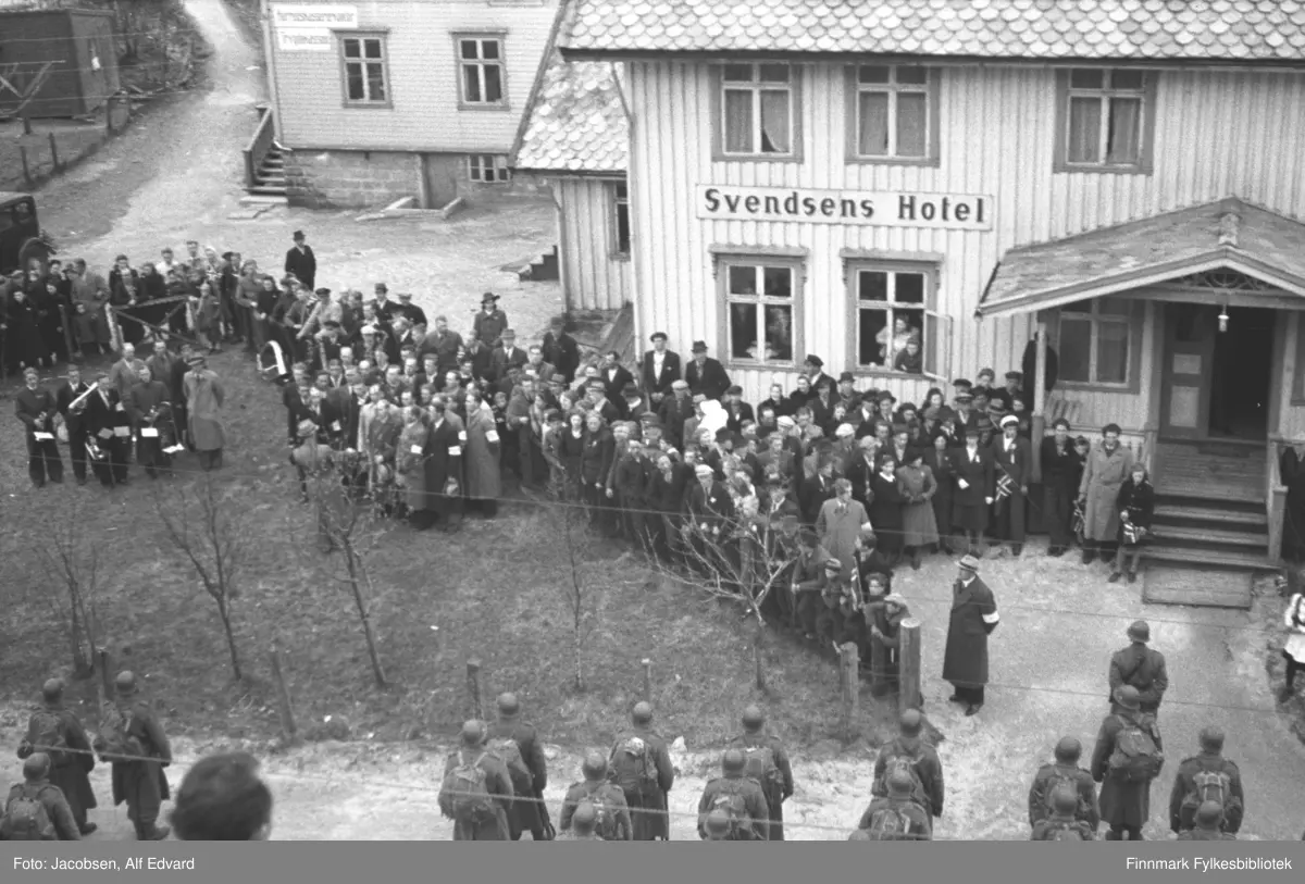 Bildet er tatt 13. mai 1945.
De norske Polititropper har ankommet med båt fra Narvik og skal sikre overgivelsen av de tyske tropper. På stedet og omegn (bl.a. Nes fort, Tjeldodden fort m.fl.). De marsjerte fra Dampskipskaia med Lødingen Musikkforening foran opp til Svendsens Hotell, hvor de ble ønsket velkommen av norske militære representanter, ordfører og lensmann. Dette bildet er tatt ved hotellet.
Vi ser også deler av folkemengden samt musikkforeningen.

Polititroppene er en betegnelse på den norske militære styrken som ble satt opp i Sverige under den andre verdenskrig, fra 1943 til 1945, med tanke på å gjøre tjeneste i Norge ved krigens slutt. 
De ble satt inn i Finnmark høsten 1944 som del av gjenerobringen og sikringen av landsdelen. De fleste av troppene ble fraktet over grensen til Norge etter 8. mai 1945 og ble satt inn flere steder i landet for å sikre freden, passe på tyske styrker og samtidig beskytte og hindre nordmenn mot å utnytte en spent og usikker fred.

Polititroppene besto av to enheter; Rikspolitiet, som fikk politiutdannelse og Reservepolitiet. Det var den siste enheten som fikk militær utdannelse og ble satt opp og trenet som en militær avdeling.