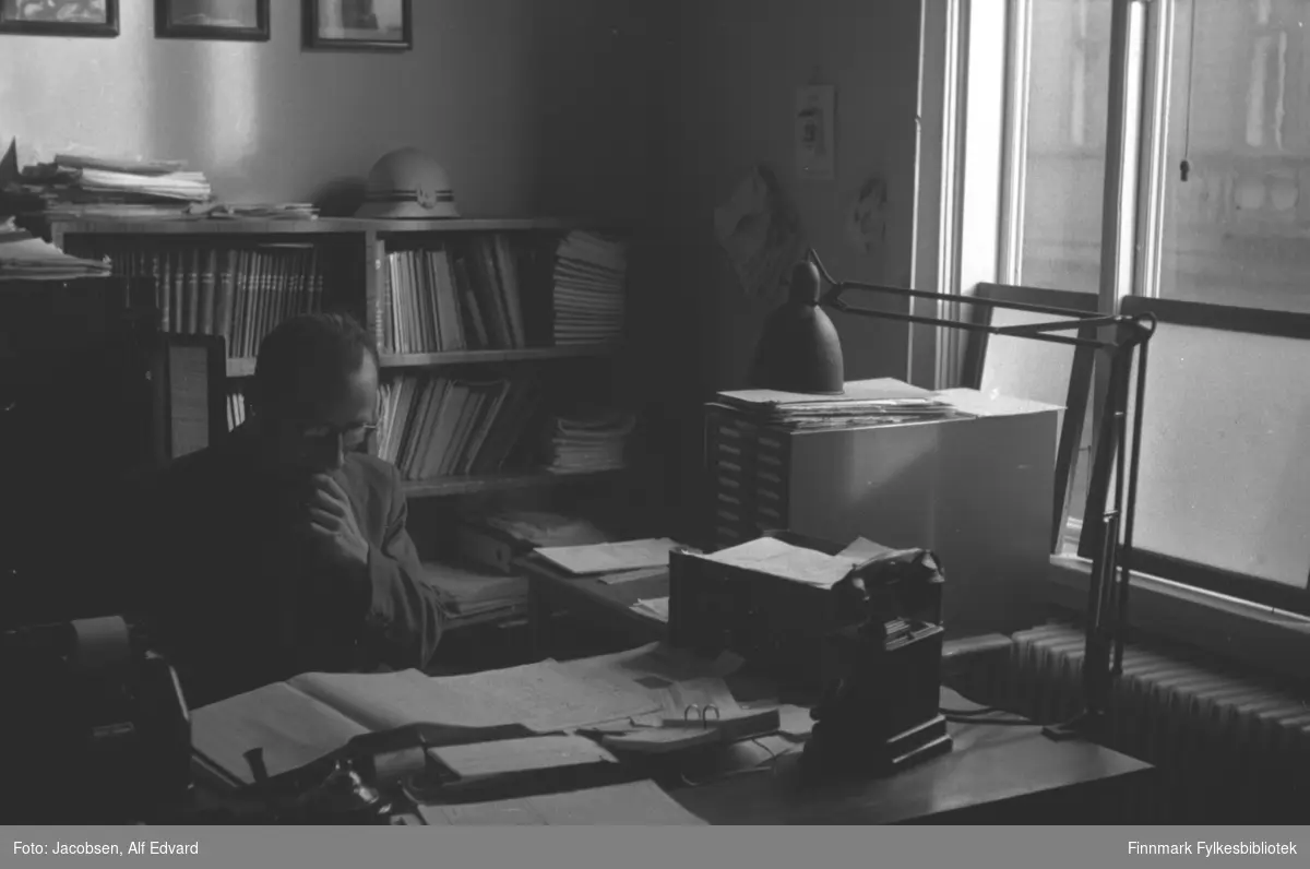 En mann sitter ved et skrivebord og leser. Iflg. informanten kan mannen være J. Carsten Knutsen som jobbet på Televerkets distriktskontor i Tromsø. Han er iført en ganske mørke jakke eller frakk og har briller på nesen. To skrivebord står i vinkel på kontoret hans. En leselampe er skrudd fast på hjørnet av det nærmeste bordet. En svart telefon står på hjørnet ved siden av lampen. Eller på skrivebordet står en liten arkivboks, en bordkalender og en del papirer. Det som står helt til venstre kan være en regnemaskin med en papirrull øverst. På vindusveggen er en stor radiator satt opp rett under vinduet. På skrivebordet ved siden av radiatoren står en større arkivboks med noe papir eller aviser oppå. Bak mannen står en bokhylle som er ganske full av bøker og hefter. Det ligger noen papirer oppå og noe hvitt/lyst som kan se ut som en hjelm. På veggen bak mannen henger tre fotografier med svarte rammer. På vindusveggen henger tre papirark, kan være tegninger. Gjennom vinduet med hvite karmer og stolpe, ses en del av et ganske stort bygg.