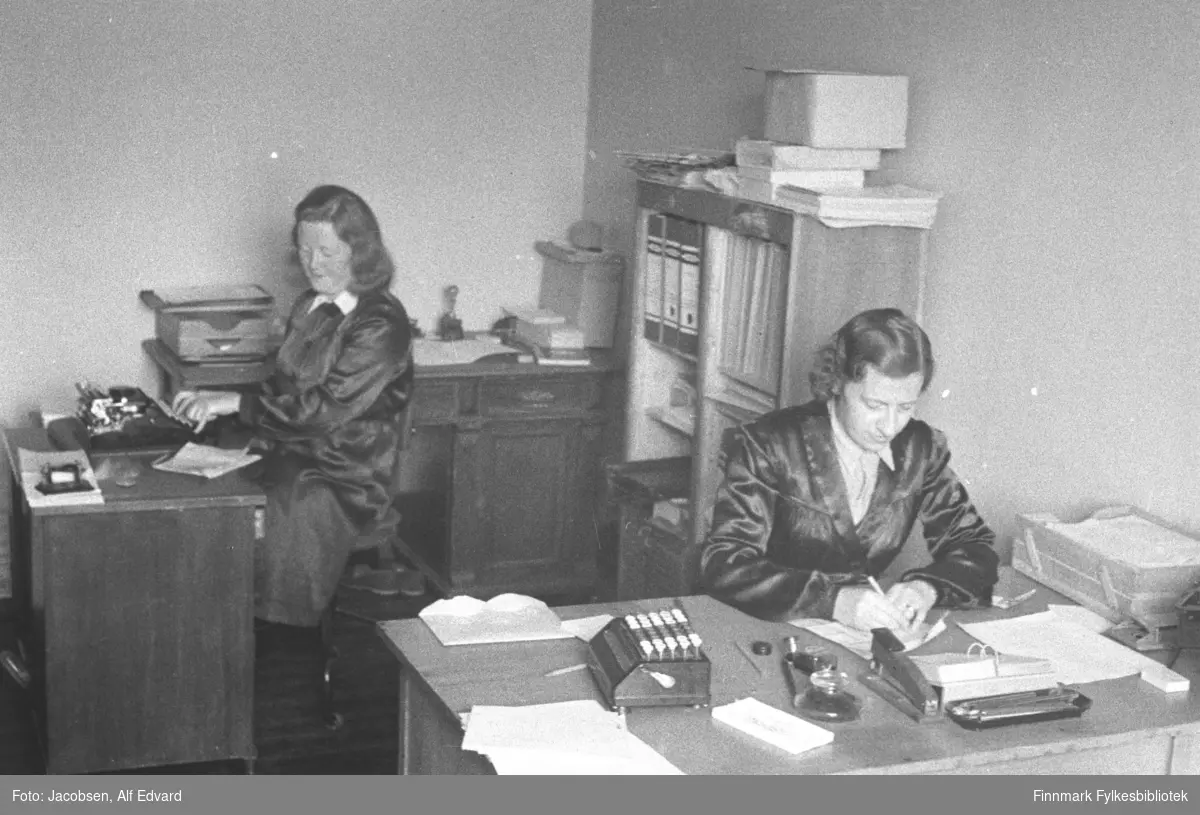 To damer sitter ved hvert sitt skrivebord. De er Randi Simonsen til venstre og Karen Iversen til høyre. Bildet er sannsynligvis fra et av Televerkets kontor i Hammerfest. Begge damene har ganske mørke frakker med lyse bluser under. På det nærmeste, ganske lyse skrivebordet, står en lys arkivkurv helt til høyre. Eller kan hullemaskin, blyantbrett, stiftemaskin, bordkalender og litt papir ses. En boks med knapper/taster som kan være en telex står til venstre på skrivebordet. Skrivebordet til venstre på bildet er ganske mørkt og oppå står en manuell skrivemaskin som damen skriver på. Litt papir ligger også på det skrivebordet. Rett bak damene står en bokhylle med flere ringpermer og hefter. Noe papir og en eske ligget oppå. Et skrivebord som står bak damen til venstre på bildet er ganske mørkt. Det har små skuffer under bordplaten og et skap på høyre side. En arkivboks står rett ved siden av damen. Lenger bort ligger noe papir og et stempel og eske ses også. Kontoret har forholdsvis lyse veggger.