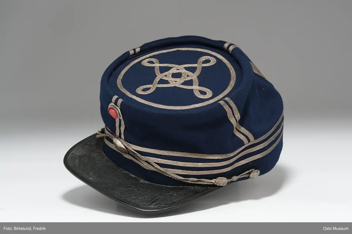 a) Våpenkjole av blått klede m. trangt liv. Kragen brodert med sølvstresser, flettede epauletter. Snorbesetning av sort silke, 3 knapperader. Fòret med sort satin. H: 97
b) Benklær av blått klede med sølvgaloner. L: 117
C) Kepi m. 3 rader sølvstresser om pullen, kokarderosett .

1875. Har tilhørt rittermester Hans Fredrik Grüner. Gave fra fru Charlotte Grüner 1913.