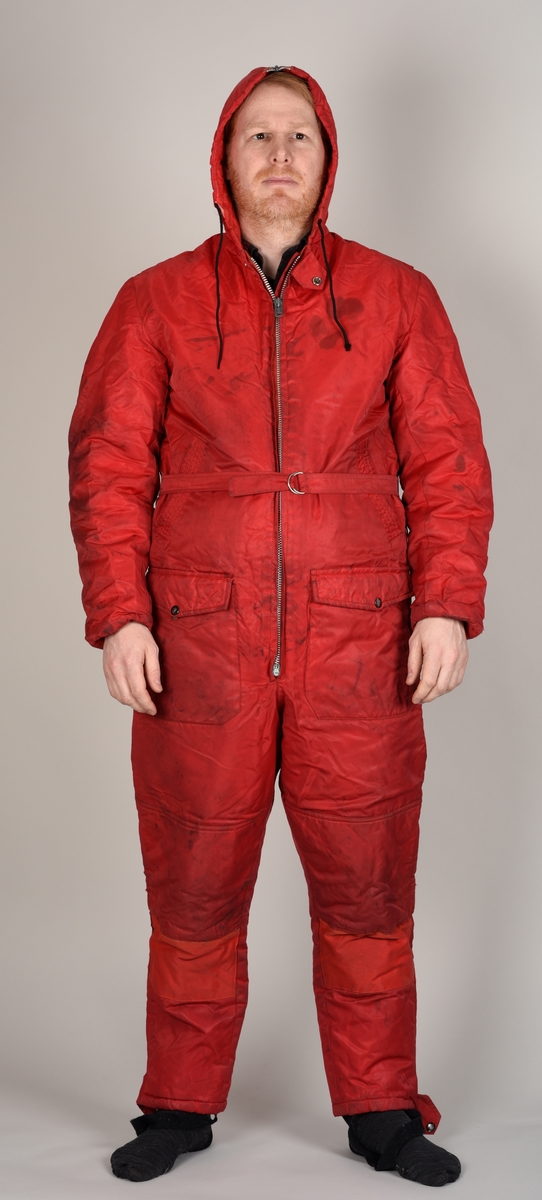 Rød dress med glidelås, hette, påsydd belte og to bukselommer.
Hetten har glidelås på oversiden.
Det er sydd på en sort stropp under hvert bein for å feste under skoene. Beltet lukkes med to halvrunde metallhemper.