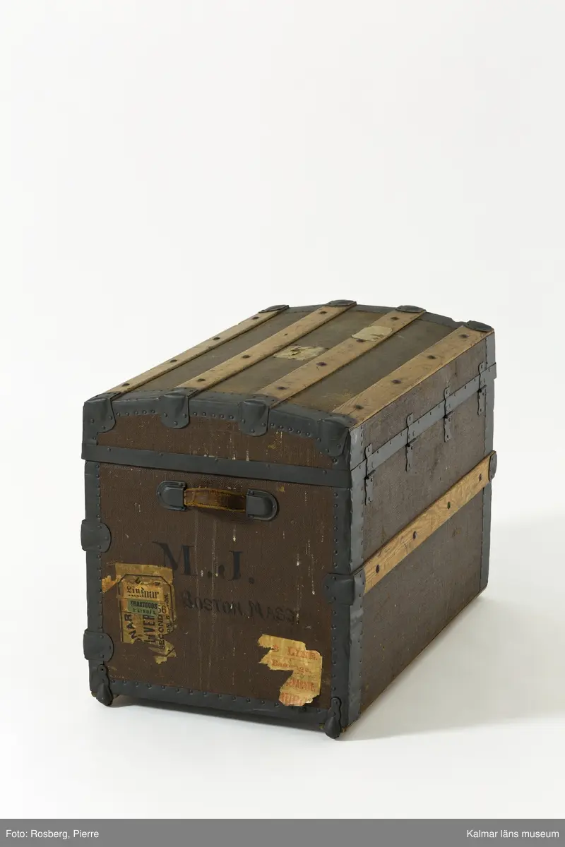 KLM 39675. Koffert, reskoffert, trä, papp, textil, läder, metall. Koffert för förvaring och resor, av trä, klädd med väv, beslag av trä och järn, lås av metall, två spännen och ett handtag av läder på varje gavel. På locket fyra beslag av trä och två på fronten. Insidan är klädd med papp eller tapet med rutigt mönster i rött, grönt och blått på gul botten. 
På lockets insida en fastklistrad tillverkaretikett med rubriken: NATHAN NEAT & CO TRUNKS and Travelling Equipage (för hela texten se foto). Text i svart på höger gavel: M.J. Boston Mass. Spår av gamla resetiketter på höger gavel. Kofferten är brunmålad. Inköpt i Amerika.