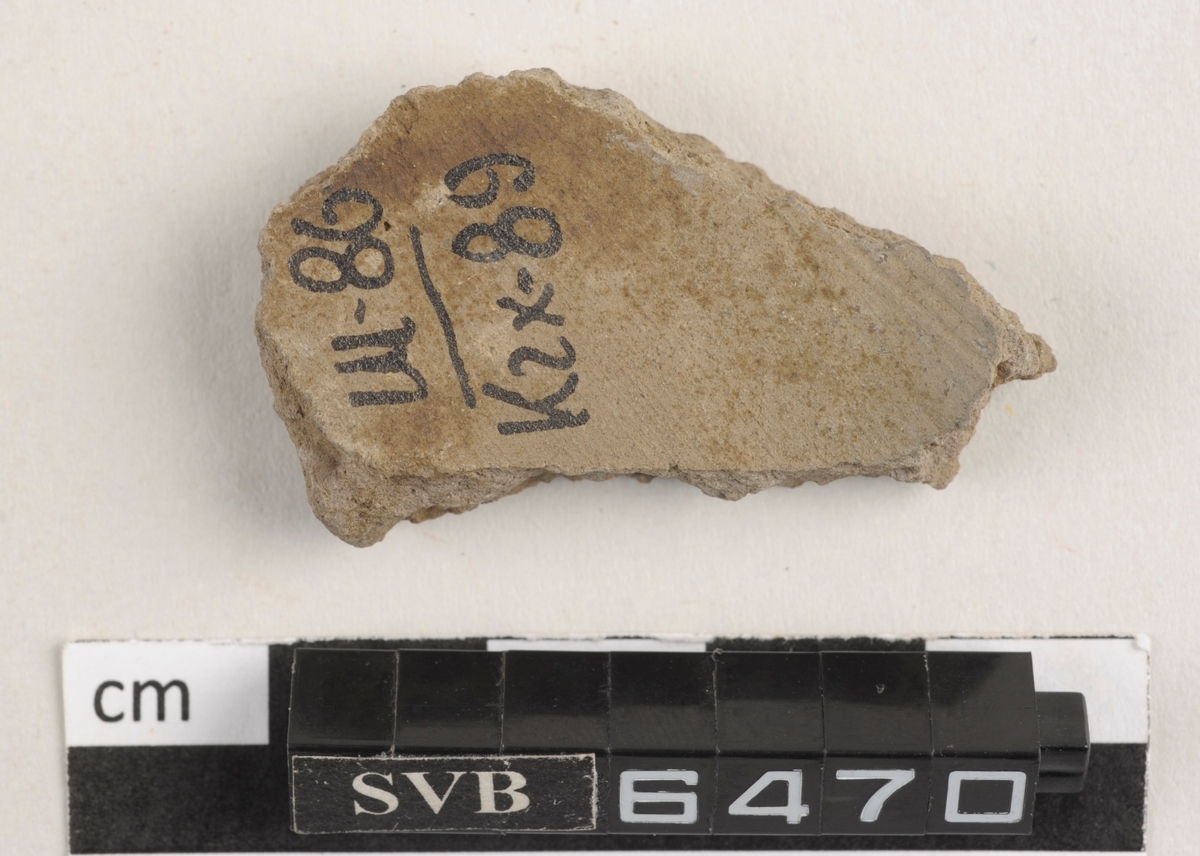 Flatt keramikkfragment med bruddkanter. Fragmentet er grått i farge.