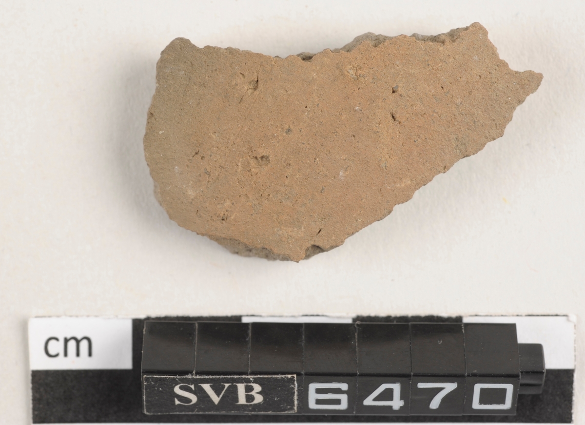 Flatt keramikkfragment med bruddkanter. Fragmentet er grått i farge.