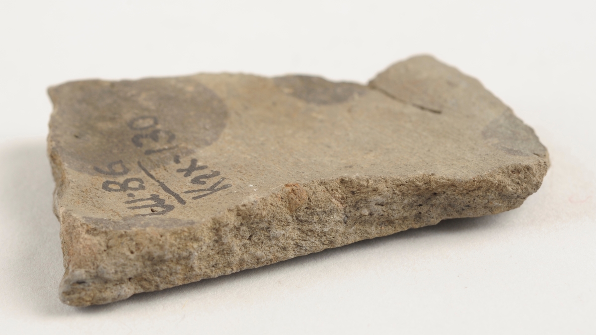 Flatt keramikkfragment med bruddkanter. Fragmentet er brunt i farge.