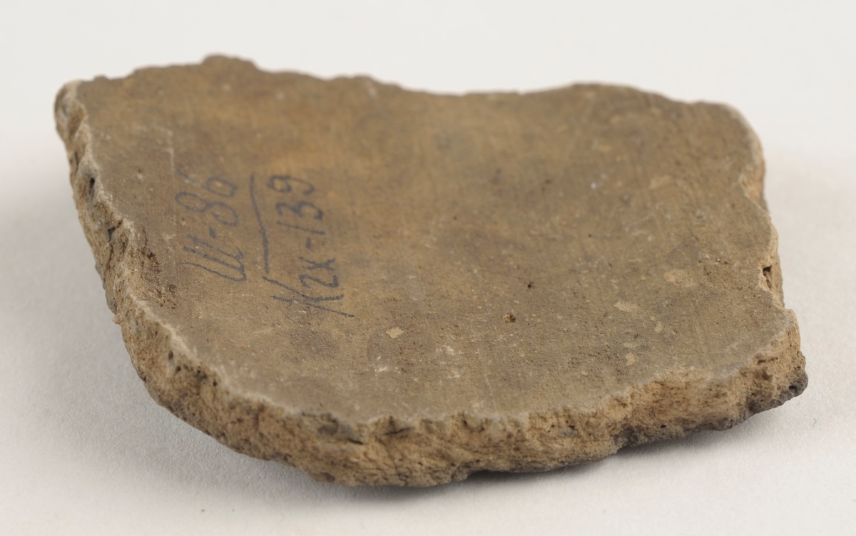 Buet keramikkfragment med bruddkanter. Fragmentet er brunt i farge.