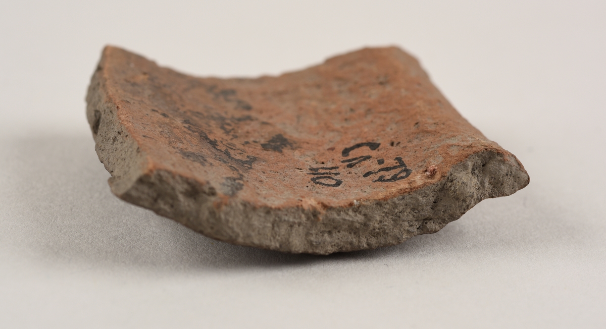 Buet keramikkfragment med bruddkanter. Fragmentet er grått i farge på utsiden og oransje i farge på innsiden.