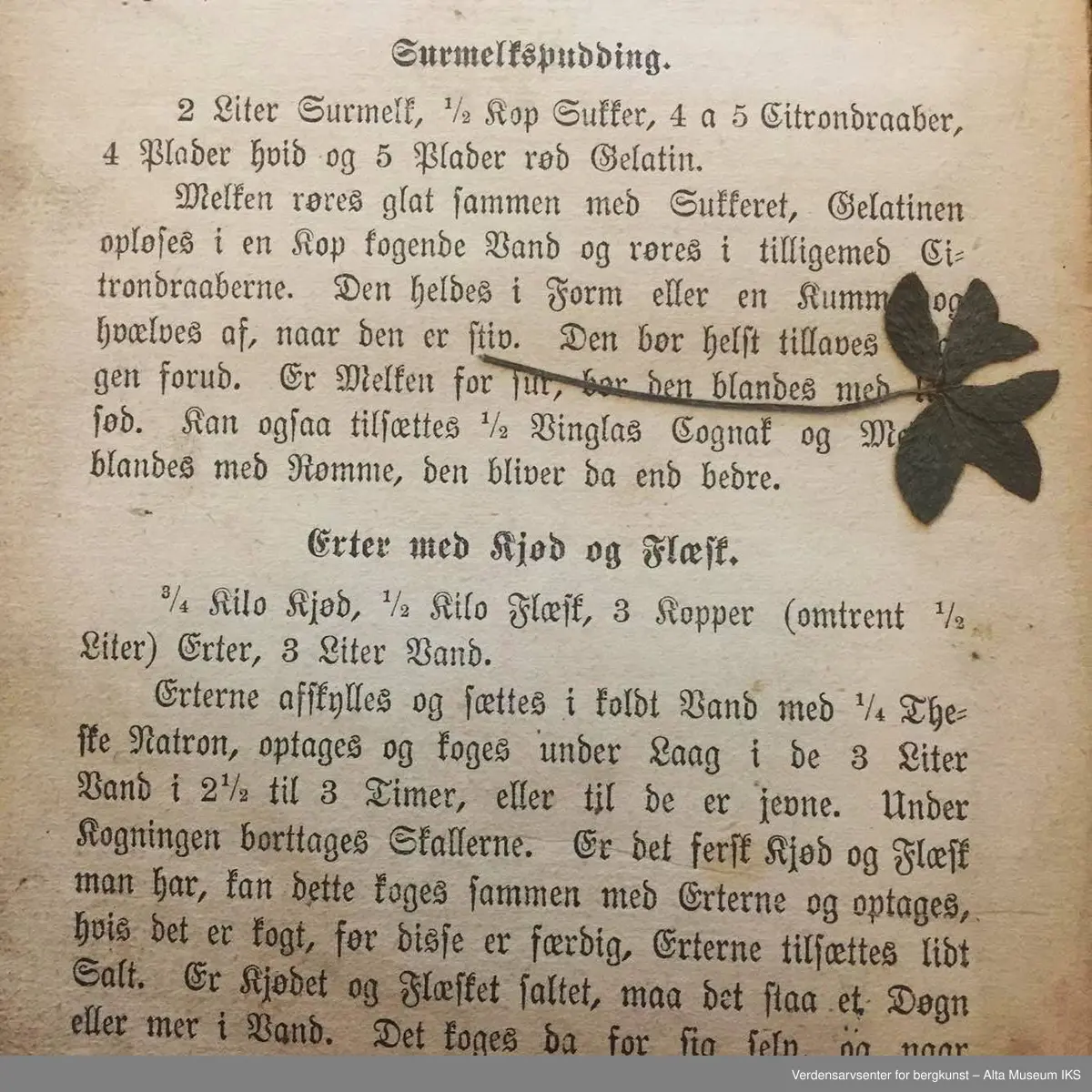 Kokebok for tarvelige husholdninger og skolekjøkkener av Antonette Baldjen. Bok med falmet og slitt omslag. Deler av boktittelen kan såvidt skimtes på bokryggen. Mellom to av bokens sider er det en presset firkløver.