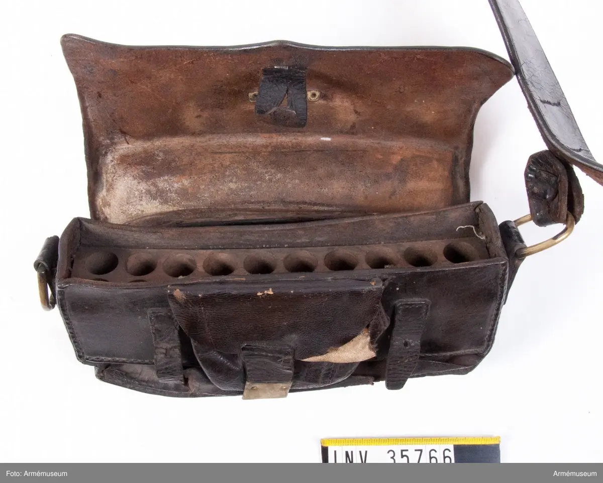 Kartuschväska av svart läder med mässingsbeslag i form av Skånes vapen. Kartuschen har en extra ficka för knallhattar och ett fack i botten som stängs med två remmar. De tillhörande spännena saknas. I kartuschen finns en träkloss med borrade hål med ca 18 mm diameter.