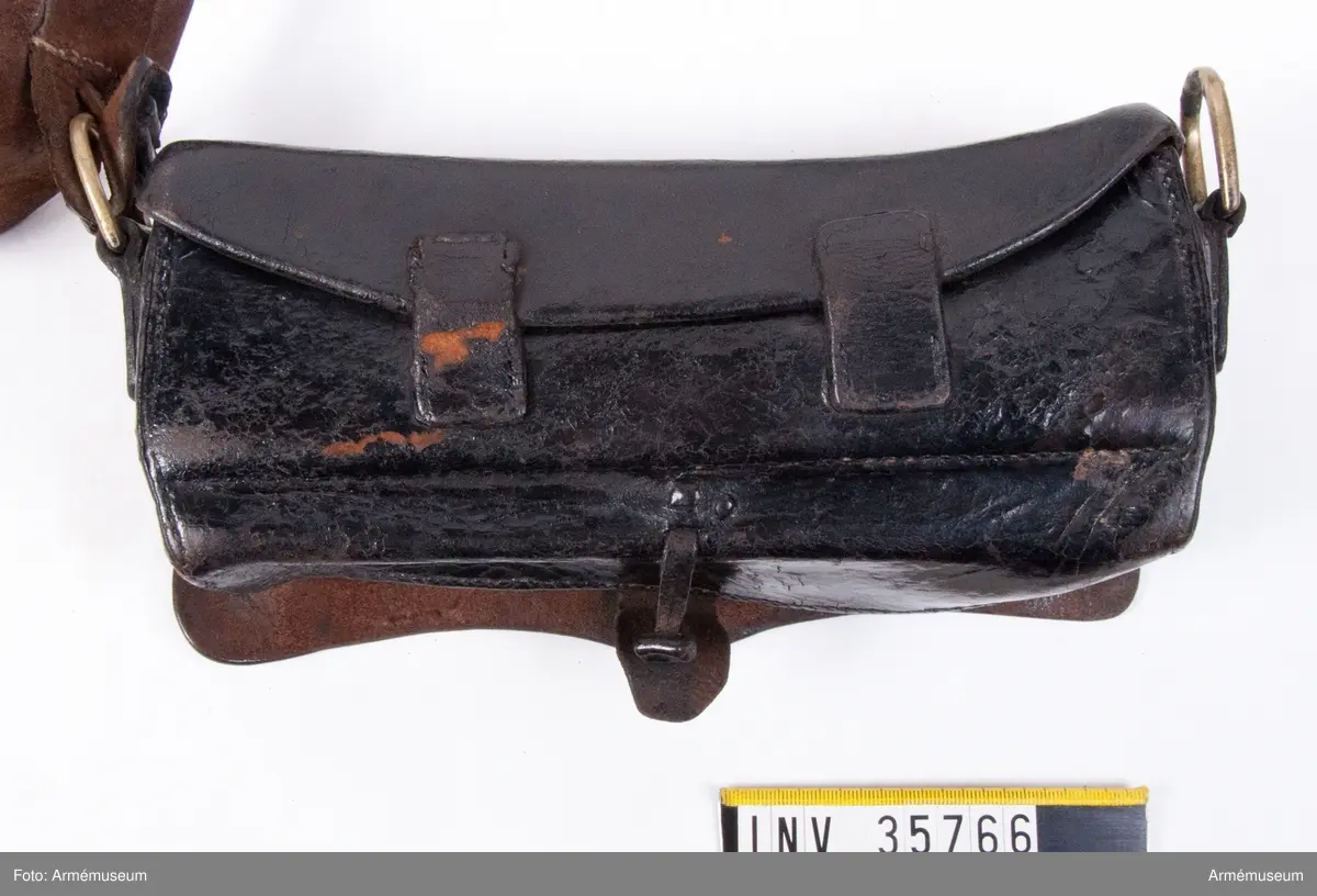 Kartuschväska av svart läder med mässingsbeslag i form av Skånes vapen. Kartuschen har en extra ficka för knallhattar och ett fack i botten som stängs med två remmar. De tillhörande spännena saknas. I kartuschen finns en träkloss med borrade hål med ca 18 mm diameter.