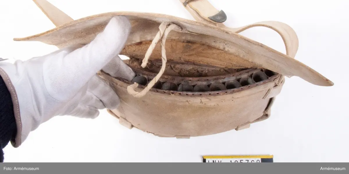 Patronväska av vitt sämskskinn med rem.
Inuti kartuchen finns en bleckplåtsinsats med rum för 12 patroner av ca 18 mm kaliber och plats för en liten oljeflaska. Under locket sitter även en extra ficka,