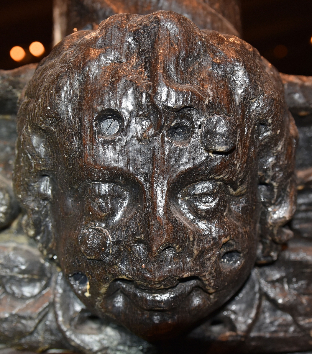 Skulpterat kerubansikte, med lockigt hår. Märken efter spikhuvuden.

Text in English: Carved sculpture of a cherub-face with curly hair. Visible marks of nail heads.