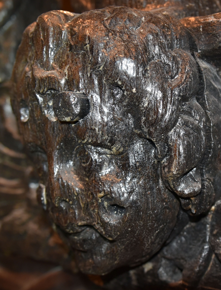 Skulpterat kerubansikte, med lockigt hår. Märken efter spikhuvuden.

Text in English: Carved sculpture of a cherub-face with curly hair. Visible marks of nail heads.