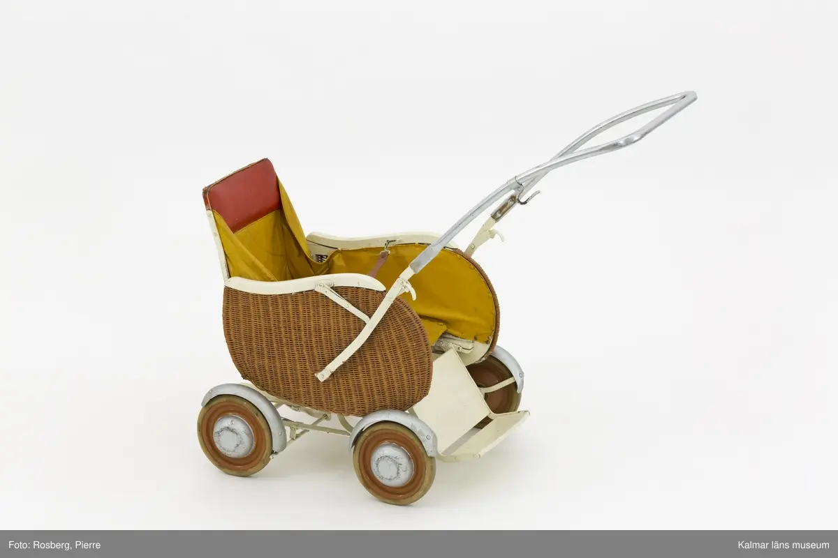 KLM 28596:2. Barnvagn, sittvagn, metall, trä, färg, textil, läder. Sittvagn för barn, ihopfällbar med stomme av järn och fyra hjul och reglerbart fotstöd av trä och järn. Sittkorgen har fastspikad klädsel av textil, målad med gul färg, och rödmålat, stoppat nackstöd. Ryggstödet är reglerbart i två lägen. Tvärs över sitsen hänger ett uppspänt läderband som fästs med hyska för barnets säkerhet. Ihopfällbart och reglerbart handtag av järn, övre delen kromat, och en krok på vänster sida. Vagnens sidor har ett korgflätat yttre lager, vitmålade sargar och silvermålade stänkskärmar och navkapslar.