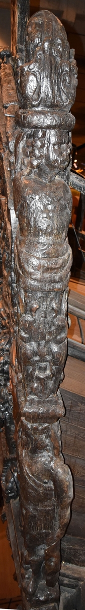 Skulptur i form av en hög, rikt snidad pelare eller stolpe, med kvadratisk genomskärning.

Tre av pelarens sidor är skulpterade medan den fjärde utgör anliggningsyta, försedd med ett antal olikformade urtag. De skulpterade sidorna är horisontellt indelade i tre eller fyra sektioner eller nivåer, avgränsade från varandra av profilerade och snedställda vulster. Nedtill avslutas skulpturen av en ornerad och på mitten insvängd formation.



Ovanför denna nedre avslutning är pelaren utformad som en reslig mansfigur, vänd mot vänster, sedd från betraktaren. Mansfiguren, som täcker tre av skulpturens fyra sidor, är iförd en romersk rustning med en bred fältbindel draperad från höger axel snett över bröstet. På huvudet med det lockiga och utslagna håret sitter en krona av öppen typ, med liljeliknande ornament. Figurens vänstra arm är böjd, medan den högra armen är sträckt utmed sidan. Handen till den högra armen saknas, men har hållit en rikt profilerad sköld, med mycket slitna och vaga konturer av en på bakbenen mot vänster gående grip med stora vingar. Skölden är snidad i ett med figuren, men med kraftiga underskärningar. Vid figurens fötter skymtar ett djur av något slag. Bakom mannens huvud tycks ett snidat textilstycke, kanske del till ett draperi, vara anbragt.



Ovanför den romerska krigaren eller riddaren har pelaren formen av en herm utan armar med långt, krusigt hår och mustaschprytt ansikte. Liksom mansfiguren täcker även hermen tre sidor av pelaren. Huvudet är vridet mot vänster. Överkroppen är bar så när som på ett flikigt plagg som täcker axlar och övre delen av bröstet. Från huvudet och runt midjan är ett tygstycke draperat. Hermens nederdel är formad till en stor maskaron med uppspärrad mun och utsträckt tunga. Maskaronen övergår upptill och utmed sidorna i yppiga bladformationer.



Pelaren avslutas upptill av ett knoppliknande ornament. Ornamentet är ornerat med akantusbladliknande bildningar samt fyra byglar som möts på toppen, där vissa spår antyder att det ursprungligen suttit ett klot eller dylikt, men som fallit bort. Eventuellt kan ornamentet utgöra en huvudbonad till hermen.

Skulpturen är kraftigt sliten.



Text in English: Sculpture in the shape of a richly carved column or pillar.

Three sides of the pillar are carved, while the fourth side comprises the attachment surface and has a number of variously-shaped indentations. The sculptured sides are divided horizontally into three or four sections separated by angled tori.



The lower section of the pillar is carved in the figure of a tall man. The figure is clad in Roman armour and has a field badge (cloak/plaid) draped across his chest at an angle from his right shoulder. On his head he wears an open crown. He has long, curly hair. The right arm is broken off, the left is at his side. The left hand is missing but once held a richly carved shield.

The shield is carved out of the same piece of wood as the figure, but deeply undercut. A carved figure is apparent on the shield. Although the shield is badly worn, it is possible to recognise one of the hind legs of a griffin with large wings. At the man's feet lies an animal of some kind.



Above the Roman warrior or knight, the pillar is carved in the shape of a herm, without arms and with long, curly hair and a moustache. Like the male figure, the herm also covers three sides of the pillar. The head is twisted to the left. The upper part of the body is bare except for a piece of cloth that covers the shoulders and upper chest. The lower part of the herm is carved as a large mask, its mouth open and tongue protruding. The sides and upper parts of the mask are carved as luxuriant leaves.



The upper part of the pillar has a ornament in shape of a knob. The ornament has a ring of acanthus leaves and four spherical loops run vertically from the acanthus leaves and meet at a point at the top. An object, for example a globe appears to have adorned the top of the ornament. This object is missing. The ornament may also be a part of the headgear of the herm.

The sculpture is severely worn.