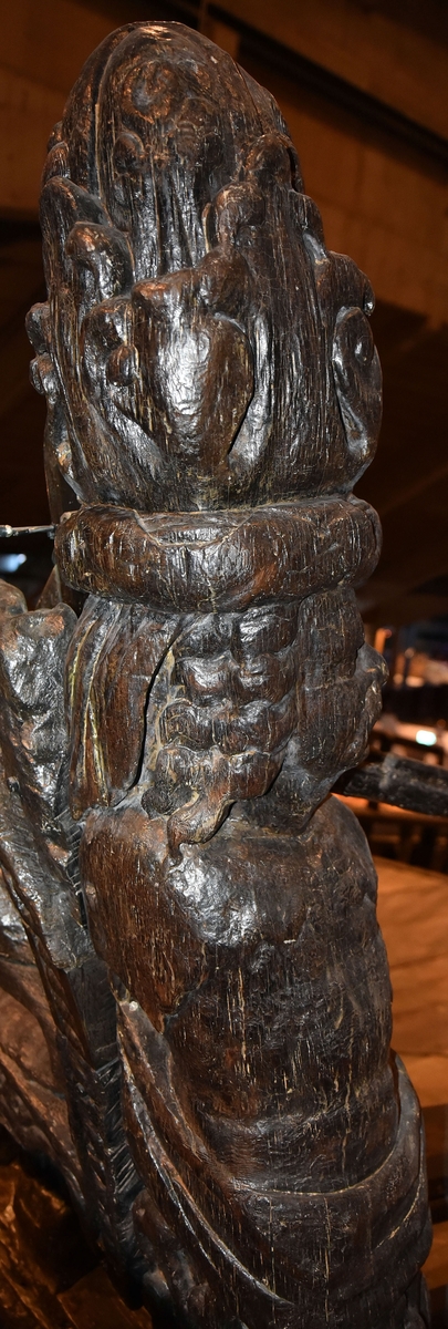Skulptur i form av en hög, rikt snidad pelare eller stolpe, med kvadratisk genomskärning.

Tre av pelarens sidor är skulpterade medan den fjärde utgör anliggningsyta, försedd med ett antal olikformade urtag. De skulpterade sidorna är horisontellt indelade i tre eller fyra sektioner eller nivåer, avgränsade från varandra av profilerade och snedställda vulster. Nedtill avslutas skulpturen av en ornerad och på mitten insvängd formation.



Ovanför denna nedre avslutning är pelaren utformad som en reslig mansfigur, vänd mot vänster, sedd från betraktaren. Mansfiguren, som täcker tre av skulpturens fyra sidor, är iförd en romersk rustning med en bred fältbindel draperad från höger axel snett över bröstet. På huvudet med det lockiga och utslagna håret sitter en krona av öppen typ, med liljeliknande ornament. Figurens vänstra arm är böjd, medan den högra armen är sträckt utmed sidan. Handen till den högra armen saknas, men har hållit en rikt profilerad sköld, med mycket slitna och vaga konturer av en på bakbenen mot vänster gående grip med stora vingar. Skölden är snidad i ett med figuren, men med kraftiga underskärningar. Vid figurens fötter skymtar ett djur av något slag. Bakom mannens huvud tycks ett snidat textilstycke, kanske del till ett draperi, vara anbragt.



Ovanför den romerska krigaren eller riddaren har pelaren formen av en herm utan armar med långt, krusigt hår och mustaschprytt ansikte. Liksom mansfiguren täcker även hermen tre sidor av pelaren. Huvudet är vridet mot vänster. Överkroppen är bar så när som på ett flikigt plagg som täcker axlar och övre delen av bröstet. Från huvudet och runt midjan är ett tygstycke draperat. Hermens nederdel är formad till en stor maskaron med uppspärrad mun och utsträckt tunga. Maskaronen övergår upptill och utmed sidorna i yppiga bladformationer.



Pelaren avslutas upptill av ett knoppliknande ornament. Ornamentet är ornerat med akantusbladliknande bildningar samt fyra byglar som möts på toppen, där vissa spår antyder att det ursprungligen suttit ett klot eller dylikt, men som fallit bort. Eventuellt kan ornamentet utgöra en huvudbonad till hermen.

Skulpturen är kraftigt sliten.



Text in English: Sculpture in the shape of a richly carved column or pillar.

Three sides of the pillar are carved, while the fourth side comprises the attachment surface and has a number of variously-shaped indentations. The sculptured sides are divided horizontally into three or four sections separated by angled tori.



The lower section of the pillar is carved in the figure of a tall man. The figure is clad in Roman armour and has a field badge (cloak/plaid) draped across his chest at an angle from his right shoulder. On his head he wears an open crown. He has long, curly hair. The right arm is broken off, the left is at his side. The left hand is missing but once held a richly carved shield.

The shield is carved out of the same piece of wood as the figure, but deeply undercut. A carved figure is apparent on the shield. Although the shield is badly worn, it is possible to recognise one of the hind legs of a griffin with large wings. At the man's feet lies an animal of some kind.



Above the Roman warrior or knight, the pillar is carved in the shape of a herm, without arms and with long, curly hair and a moustache. Like the male figure, the herm also covers three sides of the pillar. The head is twisted to the left. The upper part of the body is bare except for a piece of cloth that covers the shoulders and upper chest. The lower part of the herm is carved as a large mask, its mouth open and tongue protruding. The sides and upper parts of the mask are carved as luxuriant leaves.



The upper part of the pillar has a ornament in shape of a knob. The ornament has a ring of acanthus leaves and four spherical loops run vertically from the acanthus leaves and meet at a point at the top. An object, for example a globe appears to have adorned the top of the ornament. This object is missing. The ornament may also be a part of the headgear of the herm.

The sculpture is severely worn.