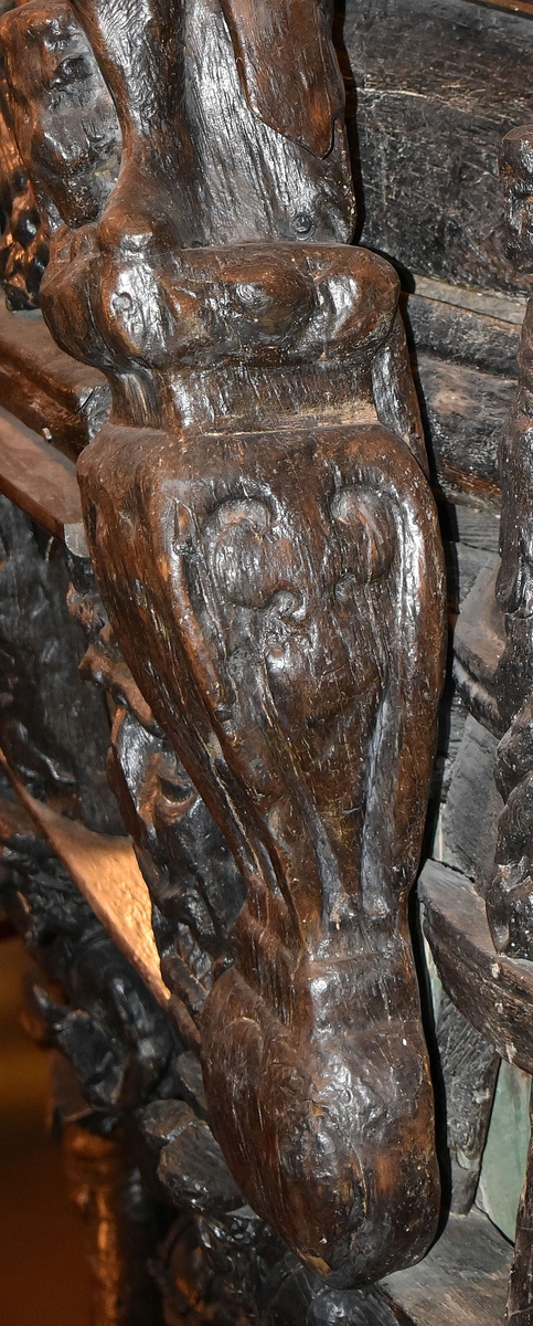 Skulptur i form av en hög, rikt snidad pelare eller stolpe, med kvadratisk genomskärning.

Tre av pelarens sidor är skulpterade medan den fjärde utgör anliggningsyta, försedd med ett antal olikformade urtag. De skulpterade sidorna är horisontellt indelade i tre eller fyra sektioner eller nivåer, avgränsade från varandra av profilerade och snedställda vulster. Nedtill avslutas skulpturen av en ornerad och på mitten insvängd formation.



Ovanför denna nedre avslutning är pelaren utformad som en reslig mansfigur, vänd mot vänster, sedd från betraktaren. Mansfiguren, som täcker tre av skulpturens fyra sidor, är iförd en romersk rustning med en bred fältbindel draperad från höger axel snett över bröstet. På huvudet med det lockiga och utslagna håret sitter en krona av öppen typ, med liljeliknande ornament. Figurens vänstra arm är böjd, medan den högra armen är sträckt utmed sidan. Handen till den högra armen saknas, men har hållit en rikt profilerad sköld, med mycket slitna och vaga konturer av en på bakbenen mot vänster gående grip med stora vingar. Skölden är snidad i ett med figuren, men med kraftiga underskärningar. Vid figurens fötter skymtar ett djur av något slag. Bakom mannens huvud tycks ett snidat textilstycke, kanske del till ett draperi, vara anbragt.



Ovanför den romerska krigaren eller riddaren har pelaren formen av en herm utan armar med långt, krusigt hår och mustaschprytt ansikte. Liksom mansfiguren täcker även hermen tre sidor av pelaren. Huvudet är vridet mot vänster. Överkroppen är bar så när som på ett flikigt plagg som täcker axlar och övre delen av bröstet. Från huvudet och runt midjan är ett tygstycke draperat. Hermens nederdel är formad till en stor maskaron med uppspärrad mun och utsträckt tunga. Maskaronen övergår upptill och utmed sidorna i yppiga bladformationer.



Pelaren avslutas upptill av ett knoppliknande ornament. Ornamentet är ornerat med akantusbladliknande bildningar samt fyra byglar som möts på toppen, där vissa spår antyder att det ursprungligen suttit ett klot eller dylikt, men som fallit bort. Eventuellt kan ornamentet utgöra en huvudbonad till hermen.

Skulpturen är kraftigt sliten.



Text in English: Sculpture in the shape of a richly carved column or pillar.

Three sides of the pillar are carved, while the fourth side comprises the attachment surface and has a number of variously-shaped indentations. The sculptured sides are divided horizontally into three or four sections separated by angled tori.



The lower section of the pillar is carved in the figure of a tall man. The figure is clad in Roman armour and has a field badge (cloak/plaid) draped across his chest at an angle from his right shoulder. On his head he wears an open crown. He has long, curly hair. The right arm is broken off, the left is at his side. The left hand is missing but once held a richly carved shield.

The shield is carved out of the same piece of wood as the figure, but deeply undercut. A carved figure is apparent on the shield. Although the shield is badly worn, it is possible to recognise one of the hind legs of a griffin with large wings. At the man's feet lies an animal of some kind.



Above the Roman warrior or knight, the pillar is carved in the shape of a herm, without arms and with long, curly hair and a moustache. Like the male figure, the herm also covers three sides of the pillar. The head is twisted to the left. The upper part of the body is bare except for a piece of cloth that covers the shoulders and upper chest. The lower part of the herm is carved as a large mask, its mouth open and tongue protruding. The sides and upper parts of the mask are carved as luxuriant leaves.



The upper part of the pillar has a ornament in shape of a knob. The ornament has a ring of acanthus leaves and four spherical loops run vertically from the acanthus leaves and meet at a point at the top. An object, for example a globe appears to have adorned the top of the ornament. This object is missing. The ornament may also be a part of the headgear of the herm.

The sculpture is severely worn.